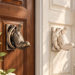 Puede incluir: Dos aldabas de puerta con forma de pájaro montadas en puertas. Una aldaba es de bronce oscuro con un acabado texturizado, y la otra es blanca con un aspecto desgastado y envejecido. Cada una presenta una base circular y una figura de pájaro.
