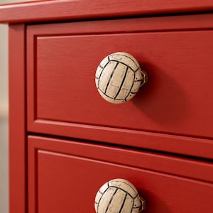 May include: Two round drawer knobs shaped like volleyballs with a cream-colored surface and black lines, attached to a bright red wooden dresser. The knobs feature a textured, weathered finish and are secured with a metallic nut on the front.