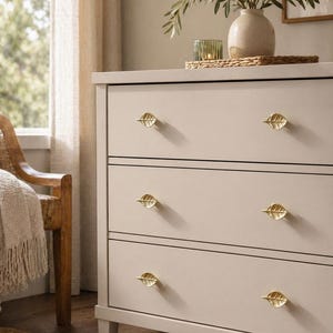 Könnte beinhalten: Eine hellbeige Kommode mit drei Schubladen, jede mit goldenen, blattförmigen Knöpfen. Die Kommode steht vor einem neutralen Hintergrund, mit einer Vase mit Grünzeug und einer Kerze, die dekorative Akzente setzen. Die Gesamtästhetik ist von schlichter Eleganz geprägt.