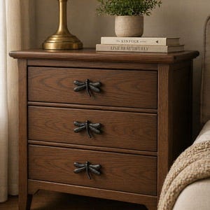 Puede incluir: Una mesita de noche de madera marrón oscuro con tres cajones, cada uno con un tirador en forma de libélula. Encima hay una lámpara con base dorada y pantalla beige, junto con libros y una pequeña planta en maceta. La mesa está junto a una cama.