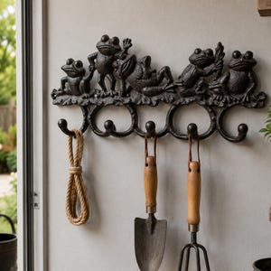 May include: A black cast iron wall hook rack featuring five sculpted frogs in various poses along the top edge. The rack includes five curved hooks, with a coiled rope, a metal hand trowel, and a metal hand cultivator hanging from them.