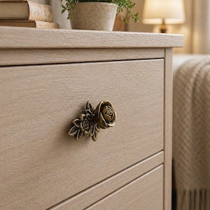 Puede incluir: Primer plano de un cajón de cómoda de madera marrón claro con un tirador decorativo en forma de rosa de bronce. El tirador presenta un diseño floral detallado. La cómoda tiene un acabado de veta de madera natural.