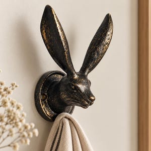 May include: A wall-mounted hook shaped like a hare head with long, upright ears. The hook features a dark, distressed black finish with metallic gold-toned highlights on the raised surfaces. A beige fabric towel hangs from the hare's neck area.