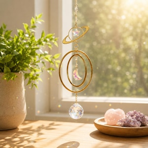 May include: A gold-coloured celestial hanging ornament featuring a planet and star, two concentric circles, and a crescent moon. A clear crystal ball hangs below. The ornament is suspended in front of a window with a plant in a vase.