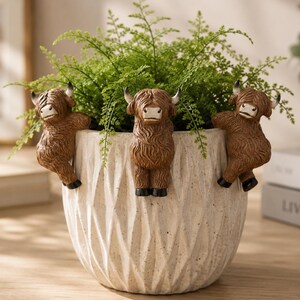 May include: A white ceramic plant pot with a geometric pattern, filled with green foliage. Three brown Highland cow ornaments are attached to the rim, adding a playful element to the home decor. The pot is approximately 20 cm in diameter.