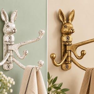 May include: Two wall-mounted hooks featuring a rabbit head design. One hook has a distressed white finish, and the other has a metallic gold finish. Each hook has three swiveling arms with rounded ends, and a beige fabric item hangs from the bottom arm.