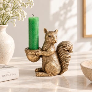 Puede incluir: Un portavelas con forma de ardilla en tono dorado con acabado texturizado. La ardilla está sentada y sostiene una copa con forma de bellota que contiene una vela de pilar verde. El diseño captura los detalles del pelaje y la cola tupida de la ardilla, proporcionando una base estable para el portavelas.