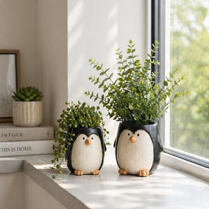 May include: Two ceramic penguin-shaped planters with black and white bodies and beige beaks and feet. The planters hold green plants, one with trailing vines. They sit on a white windowsill near a window with a view of greenery.