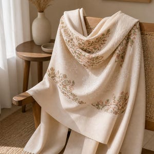 Peut inclure: Un châle crème orné d'un motif floral rose et vert, drapé sur une chaise en bois. Le châle présente une bordure à franges. L'arrière-plan comprend un vase avec des plantes séchées et une petite table d'appoint.