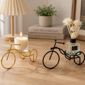 Puede incluir: Dos candelabros decorativos con forma de bicicleta. Uno es de color dorado y sostiene una vela encendida en un vaso. El otro es negro y sostiene una botella de vidrio con cañas y flores. Ambos están sobre una superficie de madera.