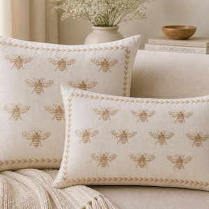 May include: Two beige linen throw pillows with embroidered bee patterns. The pillows have a decorative border with a leaf design. One pillow is rectangular, the other square. The pillows are designed for home decor.