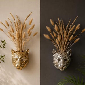 Puede incluir: Dos jarrones de pared con forma de cabeza de leopardo, uno dorado y otro plateado, cada uno lleno de espigas de trigo secas. El jarrón dorado está sobre un fondo claro, y el plateado sobre un fondo oscuro.