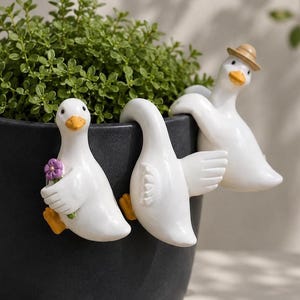 Może przedstawiać: Trzy białe ceramiczne figurki kaczek siedzące na czarnym doniczce wypełnionej zielonymi roślinami. Jedna kaczka trzyma fioletowy kwiat, a druga ma na sobie mały słomkowy kapelusz. Kaczki mają pomarańczowe dzioby i stopy.