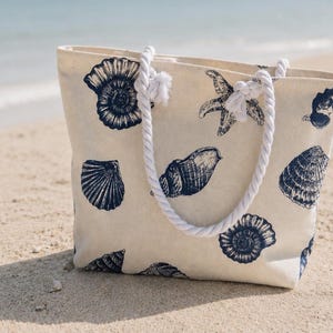 May include: A cream-colored tote bag with navy blue seashell designs. The bag has white rope handles and is sitting on a sandy beach. The bag features various seashell illustrations, including starfish and spiral shells.