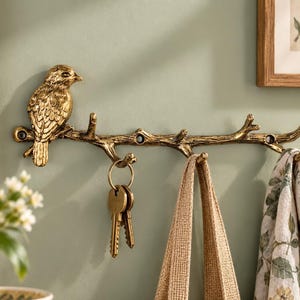 May include: A gold-toned metal wall hook shaped like a tree branch with a bird perched on the left side. The branch features multiple hooks for hanging items, with a set of keys, a beige fabric strap, and a patterned cloth hanging from it.