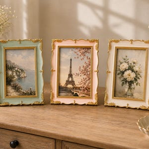 May include: Three rectangular picture frames with ornate gold-toned edges and pastel-coloured borders in light blue, soft pink, and cream. Each frame displays a different scenic print, including a coastal landscape, the Eiffel Tower, and flowers.