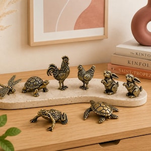 May include: A collection of eight small, antique-style brass animal figurines including a dragonfly, two turtles, a rooster, a hen, two rabbits, and a frog. These detailed metal sculptures are arranged on a smooth, light-coloured, kidney-shaped stone tray.