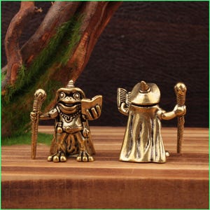 May include: Two brass frog figurines, one holding a staff and a book, the other with a staff and a cloak. The figurines are on a wooden surface with a dark brown background and a tree branch.
