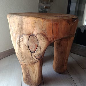 Può includere: Uno sgabello piccolo ricavato da un unico pezzo di legno marrone chiaro, caratterizzato da una seduta circolare piatta e tre gambe spesse e robuste. È visibile la venatura naturale del legno, incluso un nodo scuro sul corpo principale.