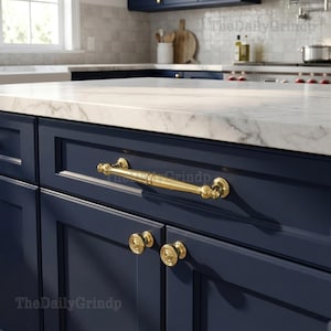 May include: Close-up of a navy blue kitchen cabinet with gold hardware. The cabinet features a long, curved handle and two smaller round knobs. A white marble countertop is visible above the cabinet.