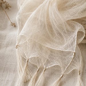 May include: A sheer, off-white scarf featuring a delicate, web-like fibrous texture throughout the fabric. The edges of the scarf are finished with a series of evenly spaced, thin, matching off-white tassels that hang from small, knotted triangular sections.