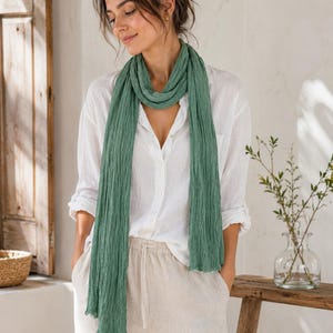 May include: A long, sage green scarf with a crinkled, textured fabric finish. The scarf is draped loosely around the neck, hanging down the front in two long, vertical panels with raw, unfinished edges at the bottom hem of the accessory.