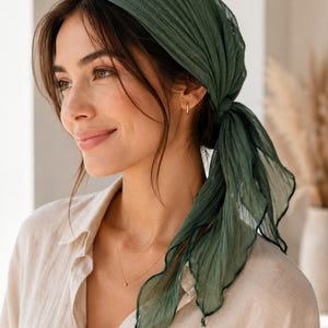 Op de afbeelding: Een donker bosgroene hoofddoek gemaakt van een lichtgewicht, semi-transparante, gekreukte stof. De vierkante sjaal is diagonaal gevouwen en stevig aan de achterkant van het hoofd geknoopt, met een delicate golvende zoom langs de randen.
