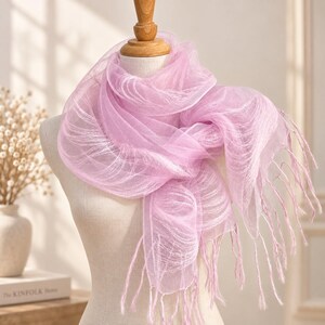 May include: A light lavender scarf made of a sheer, translucent fabric with a fine mesh texture. The scarf is draped around a mannequin neck and features long, thin, matching lavender fringe tassels hanging from the ends of the material.
