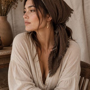 May include: A dark brown headscarf featuring thin, vertical white stitched lines and frayed edges, tied securely around the head. The fabric has a textured appearance, and the scarf is worn with a light beige long-sleeved button-down shirt.