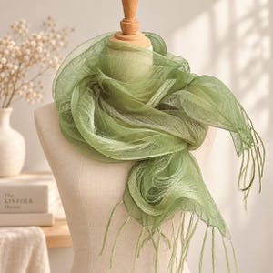 May include: A sage green scarf made of a sheer, lightweight, and translucent fabric with a delicate, airy texture. The scarf features a soft, flowing drape around the neck area, finished with thin, dangling fringe tassels at the ends of the fabric.