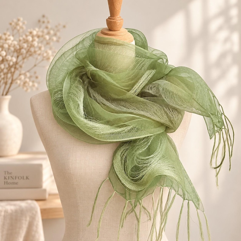 May include: A sage green scarf made of a sheer, lightweight, and translucent fabric with a delicate, airy texture. The scarf features a soft, flowing drape around the neck area, finished with thin, dangling fringe tassels at the ends of the fabric.