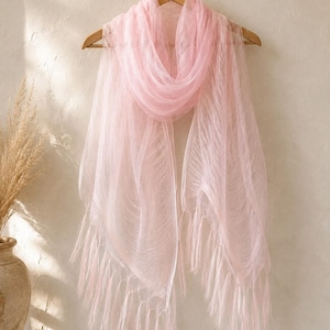 May include: A light pink, sheer, rectangular scarf with a delicate, crinkled texture and long, flowing fringe along the bottom edges. The scarf is draped over a wooden hanger, showcasing its translucent fabric and soft, pale pink color.