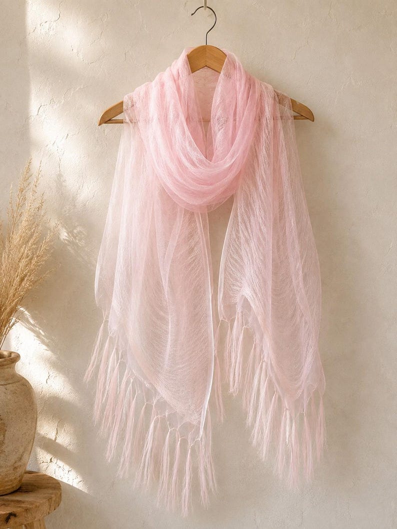 May include: A light pink, sheer, rectangular scarf with a delicate, crinkled texture and long, flowing fringe along the bottom edges. The scarf is draped over a wooden hanger, showcasing its translucent fabric and soft, pale pink color.