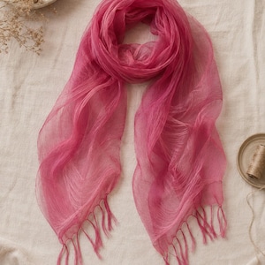 May include: A sheer, fuchsia-colored scarf made of a lightweight, translucent fabric. The scarf features a soft, draped texture and is finished with delicate, matching fringe tassels along both of the bottom edges, creating a flowing appearance.