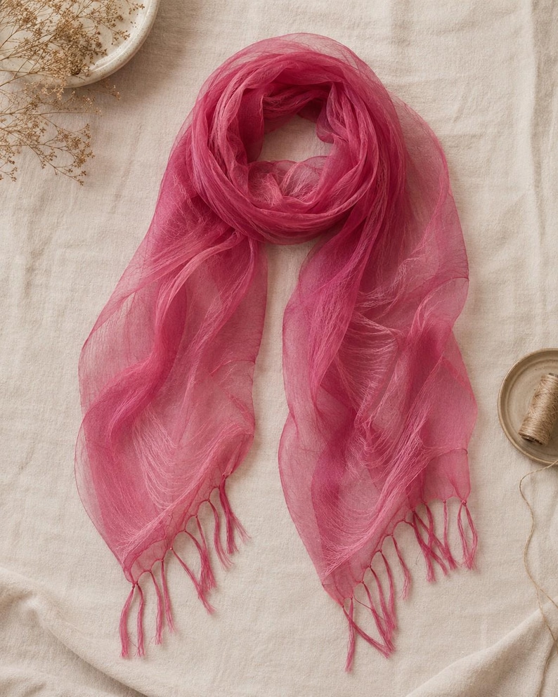 May include: A sheer, fuchsia-colored scarf made of a lightweight, translucent fabric. The scarf features a soft, draped texture and is finished with delicate, matching fringe tassels along both of the bottom edges, creating a flowing appearance.