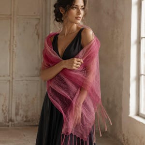 May include: A sheer, magenta-colored shawl with a delicate, web-like texture and long, thin fringe along the edges. The shawl is draped over a simple, sleeveless black dress with a deep V-neckline, creating a layered and elegant appearance.