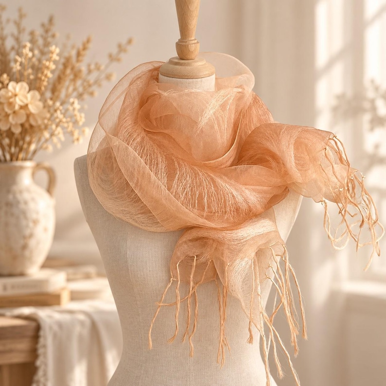 May include: A sheer, peach-colored scarf with a delicate, translucent texture draped around a mannequin. The scarf features long, thin fringe detailing along the edges, creating a soft, layered appearance with light filtering through the fabric.