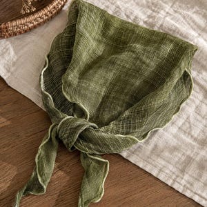 May include: A square olive green scarf with a visible cross-hatched weave pattern and a light green lettuce-edge trim. The scarf is tied in a simple knot, showcasing the lightweight, semi-sheer fabric texture and the wavy, decorative border.