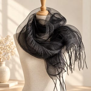 May include: A sheer black scarf with a delicate, translucent texture draped around a mannequin. The scarf features a layered, voluminous design with fine, wispy fringe detailing along the edges, creating a soft and airy appearance.