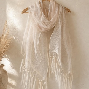 May include: A lightweight, semi-sheer scarf in a soft beige hue, featuring a delicate, fine-textured weave throughout the fabric. The scarf is draped over a wooden hanger and is finished with long, thin, matching fringe tassels along the bottom edge.