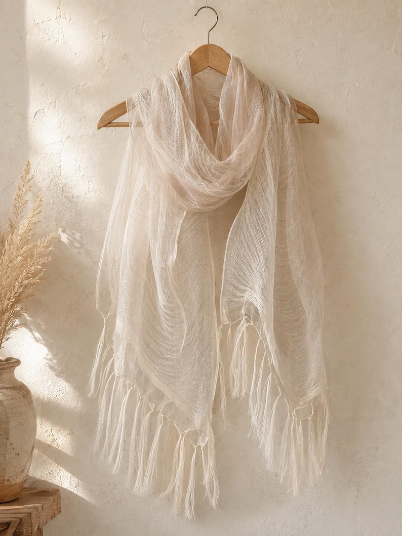 May include: A lightweight, semi-sheer scarf in a soft beige hue, featuring a delicate, fine-textured weave throughout the fabric. The scarf is draped over a wooden hanger and is finished with long, thin, matching fringe tassels along the bottom edge.