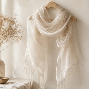 May include: A lightweight, semi-sheer off-white scarf with a delicate, gauzy texture. The scarf is draped over a wooden hanger and features long, thin fringe detailing along the bottom edges, creating a soft, flowing appearance in a neutral tone.