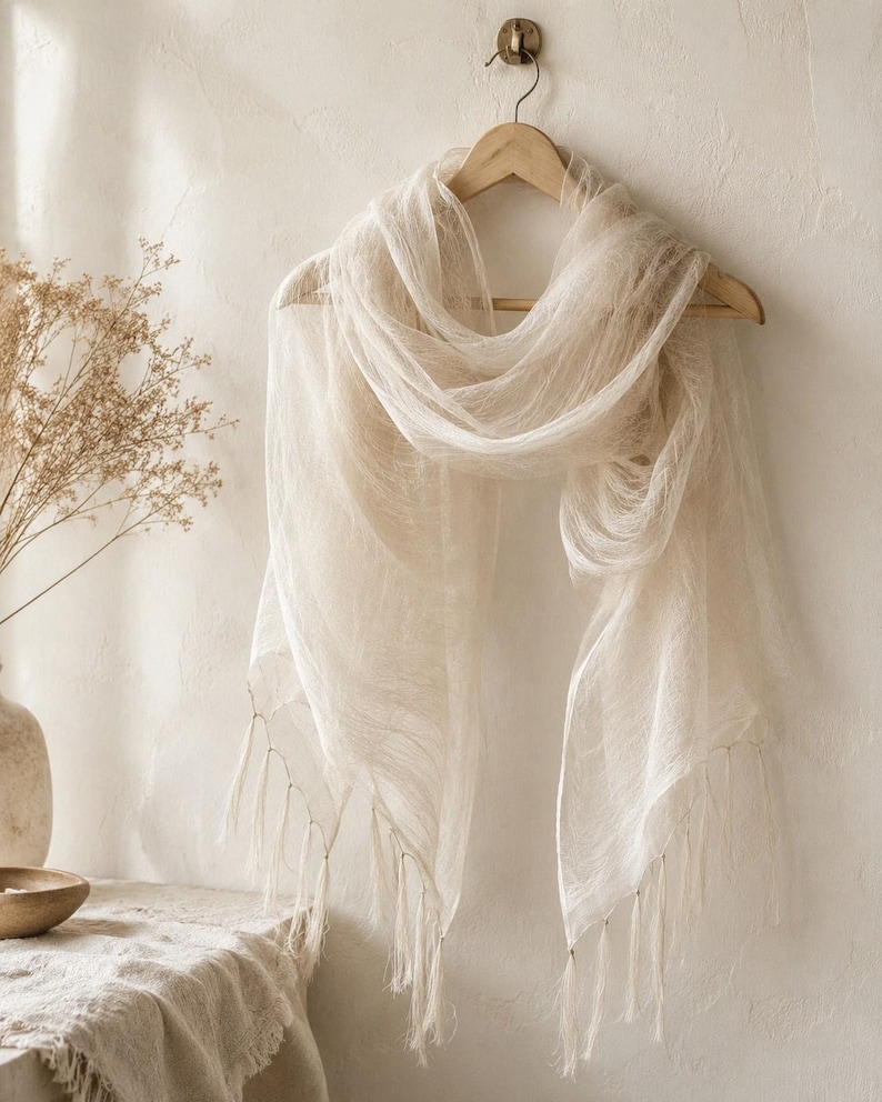 May include: A lightweight, semi-sheer off-white scarf with a delicate, gauzy texture. The scarf is draped over a wooden hanger and features long, thin fringe detailing along the bottom edges, creating a soft, flowing appearance in a neutral tone.