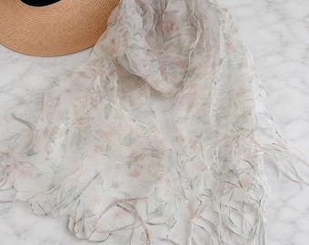 May include: A sheer, lightweight scarf featuring a delicate floral pattern in soft shades of pink and green on a white background, finished with frayed edges, positioned next to a tan straw hat with a wide brim and a black ribbon band.