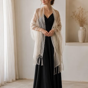 May include: A sheer, champagne-colored shawl with a delicate, semi-transparent texture and fringe detailing along the bottom edges, draped over a solid black, sleeveless floor-length dress with a V-neckline and a fitted bodice.