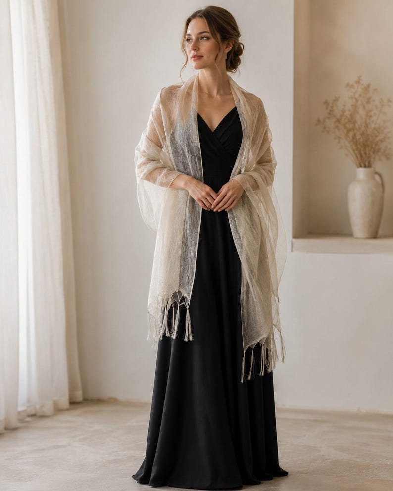 May include: A sheer, champagne-colored shawl with a delicate, semi-transparent texture and fringe detailing along the bottom edges, draped over a solid black, sleeveless floor-length dress with a V-neckline and a fitted bodice.