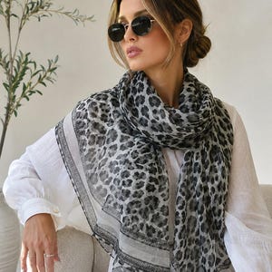 May include: A lightweight scarf featuring a grey and black leopard print pattern with a solid white border and a thin black striped edge. The fabric has a semi-sheer texture and is draped around the neck over a plain white long-sleeved shirt.