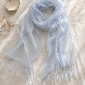 May include: A light blue, semi-sheer scarf with a delicate, airy texture and fine fringe detailing along the edges. The fabric appears lightweight and slightly translucent, featuring a soft, cool-toned color palette throughout the entire piece.