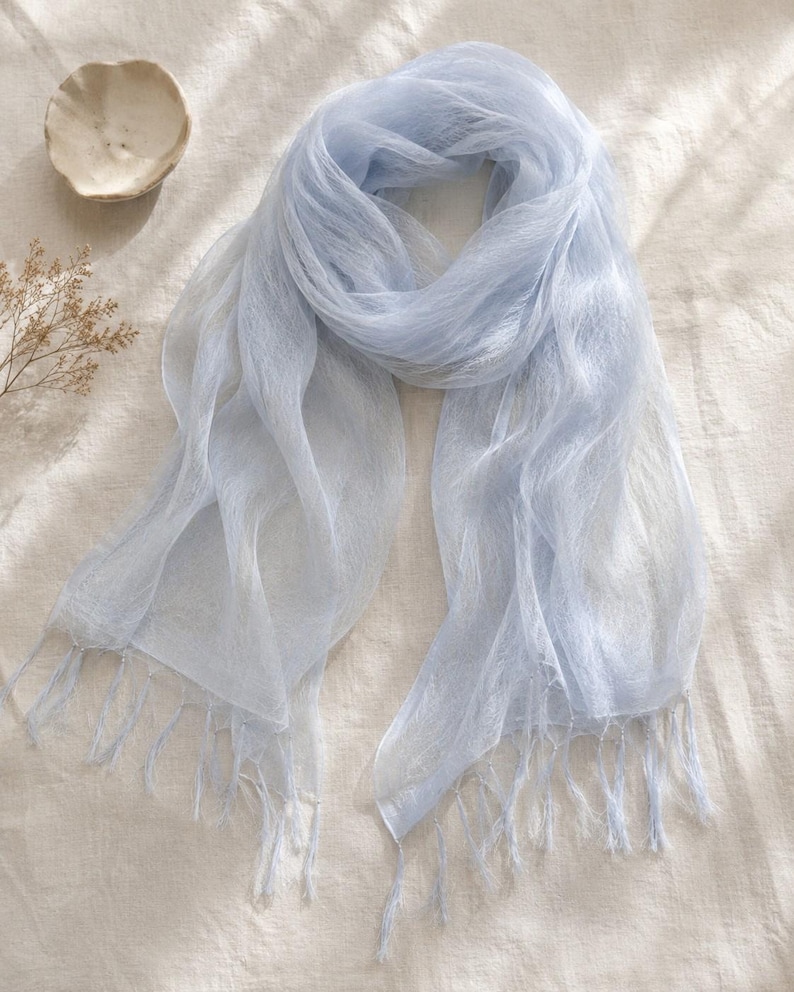May include: A light blue, semi-sheer scarf with a delicate, airy texture and fine fringe detailing along the edges. The fabric appears lightweight and slightly translucent, featuring a soft, cool-toned color palette throughout the entire piece.