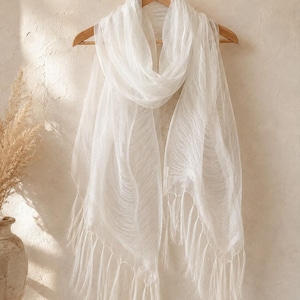 May include: A lightweight, semi-sheer white scarf with a delicate, crinkled texture and long, flowing fringe along the bottom edges. The scarf is draped over a wooden hanger, showcasing its soft, airy fabric and elongated rectangular shape.
